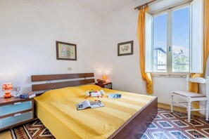 Een hotelkamer met een bed, een stoel, een nachtkastje, een raam met gordijnen en een muur met twee ingekaderde foto’s.