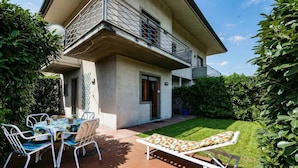 Una casa de dos pisos con un balcón, un patio con mesa y sillas, y un jardín.
