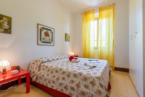 A bedroom with a bed, a nightstand, a lamp, and a window with yellow curtains. - La Rosa - Focette (Pietrasanta)