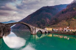 A stone bridge over a calm river, with buildings and hills in the background.