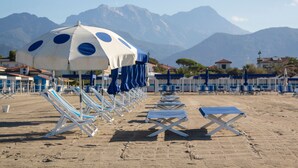 Una playa con filas de sillas de playa y sombrillas, arena dorada y montañas al fondo.