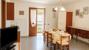 Un comedor con una mesa dispuesta para cuatro, un televisor sobre un mueble bajo y una zona de cocina con armarios y un refrigerador.