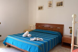 Un lit bien fait, avec une couvre-lit à rayures, une serviette enroulée et un magazine ouvert posé sur le lit. On y trouve une table de chevet en bois avec une lampe, ainsi qu’une petite table sur laquelle est posé un objet coloré.