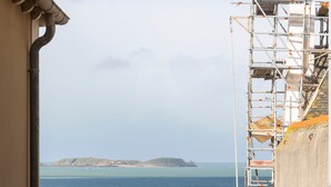 Une vue sur la côte, prise depuis une fenêtre où une tasse est posée sur le rebord, un bâtiment en construction avec des échafaudages, et une côte rocheuse.