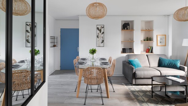 Un salon moderne avec un canapé, une table basse et une table à manger avec des chaises. Des objets décoratifs sont disposés sur des étagères, et un grand miroir reflète l’ensemble de la pièce.