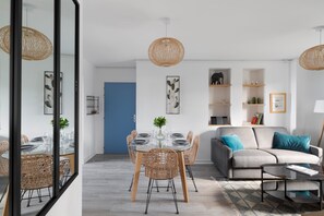 Una sala moderna con sofá, mesa de centro y mesa de comedor con sillas. Hay elementos decorativos en estanterías y un espejo grande que refleja la habitación.