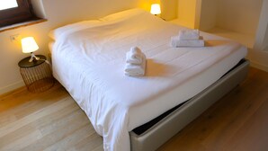 Ein ordentlich bezogenes Bett mit weißen Leinentüchern und zwei darauf gefalteten Handtüchern. Ein kleiner Tisch mit einer Lampe steht neben dem Bett.