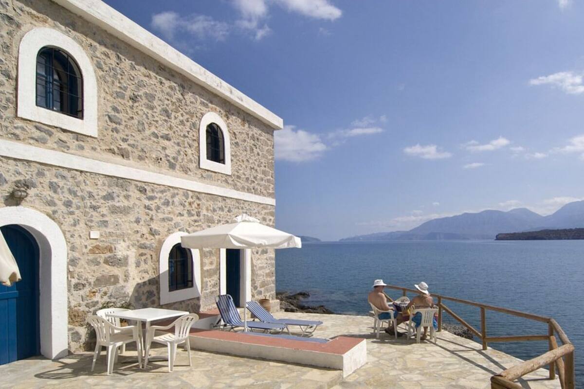 A stone building with a terrace overlooking a body of water, featuring outdoor seating and a view of distant mountains.