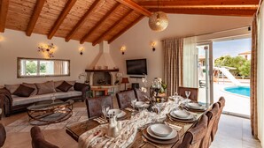 Uma sala de jantar com uma mesa de madeira posta para seis lugares, um sofá e uma televisão ao fundo.