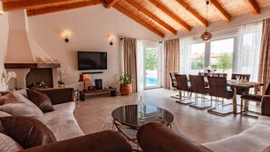 Uma sala de estar ampla com teto de madeira, lareira, televisão de tela plana, mesa de jantar com cadeiras e uma grande janela com cortinas.