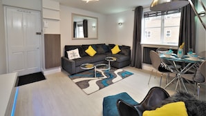 En moderne stue med en grå sofa, et sofabord, et spiseområde med bord og stole, samt et stort vindue med gardiner.