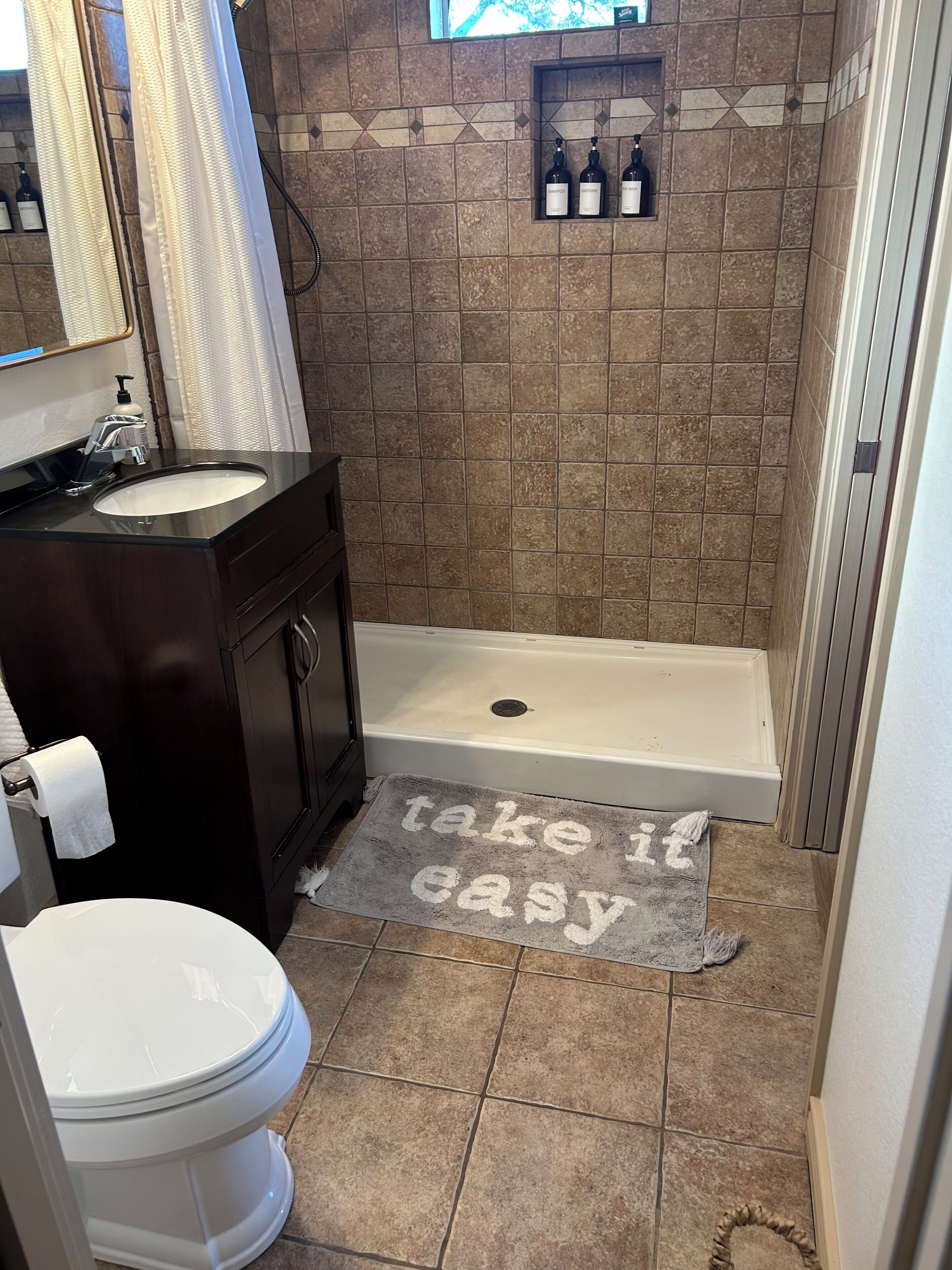 A bathroom with a dark wood vanity, a white sink, a toilet, and a bathtub. There is a shower curtain, a towel rack, and a rug with the text "take it easy."