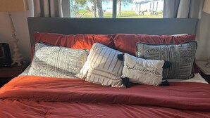 Un lit recouvert d’une couverture rouge et agrémenté de plusieurs coussins à motifs, une table de chevet avec une lampe, et une vue sur des arbres et une pelouse, visible par la fenêtre.