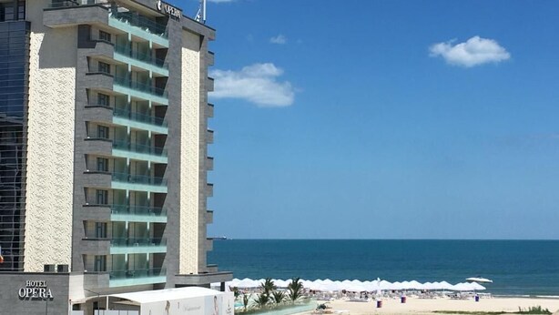 Ein Hotel namens 'Hotel Opera' mit modernem Design, nahe einem Strand mit Sonnenschirmen und freiem Blick auf den Ozean.