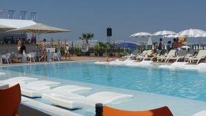 Ein Poolbereich mit Liegestühlen und Sonnenschirmen, ein Barbereich mit Sitzgelegenheiten und eine Bühne mit Lautsprechern.