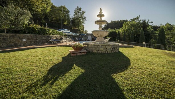 A fountain with a statue on top, surrounded by a stone base, with a lawn and trees in the background.