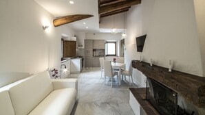 A modern living room with a white sofa, a dining area with a white table and chairs, a kitchen with wooden cabinets, and a fireplace.