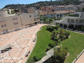A courtyard with a geometric metal structure, surrounded by buildings with balconies and a grassy area with shrubs.