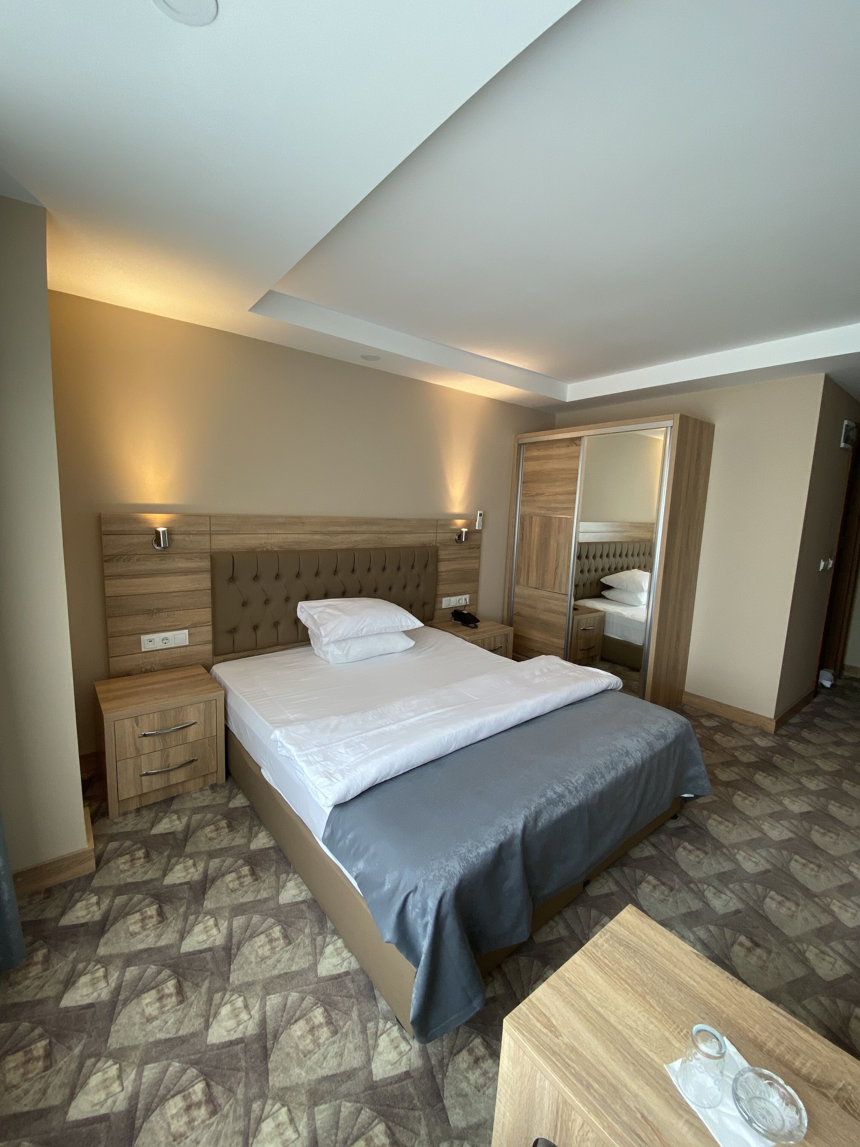 Hotelkamer met een groot bed, een houten hoofdbord, nachtkastjes, een garderobe en een klein houten tafeltje met glazen.