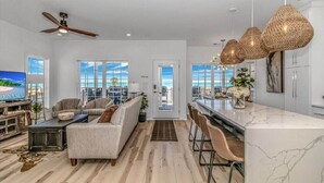 A spacious living room with a large window, a ceiling fan, and a kitchen area with a marble countertop and wicker pendant lights.