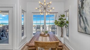 A dining area with a wooden table, chairs, a chandelier, and a view of the beach.