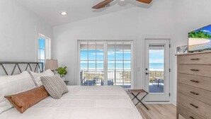A bedroom with a large bed, a ceiling fan, a TV, and a balcony with a view of the beach.