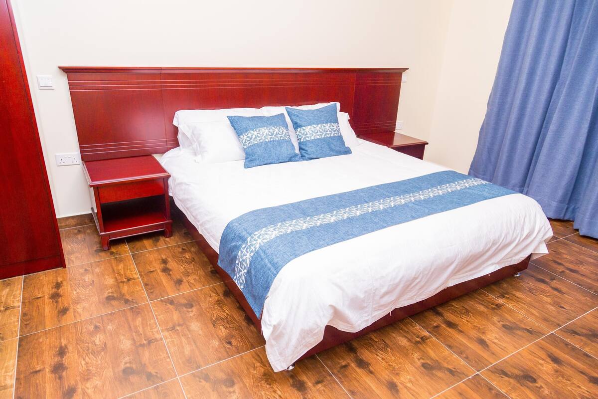 Una cama bien hecha con una colcha blanca y azul, un cabecero de madera y una mesita de noche de madera con acabado rojo.