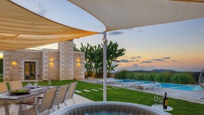 A hot tub with a pool and lounge chairs, a table with a bowl of fruit, and a stone building with lights.