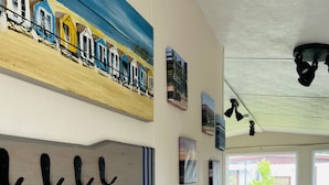 Um corredor com uma placa de madeira com a inscrição 'MAR, SOL, AREIA', uma pintura de casas de praia e uma área de jantar com mesa e cadeiras.
