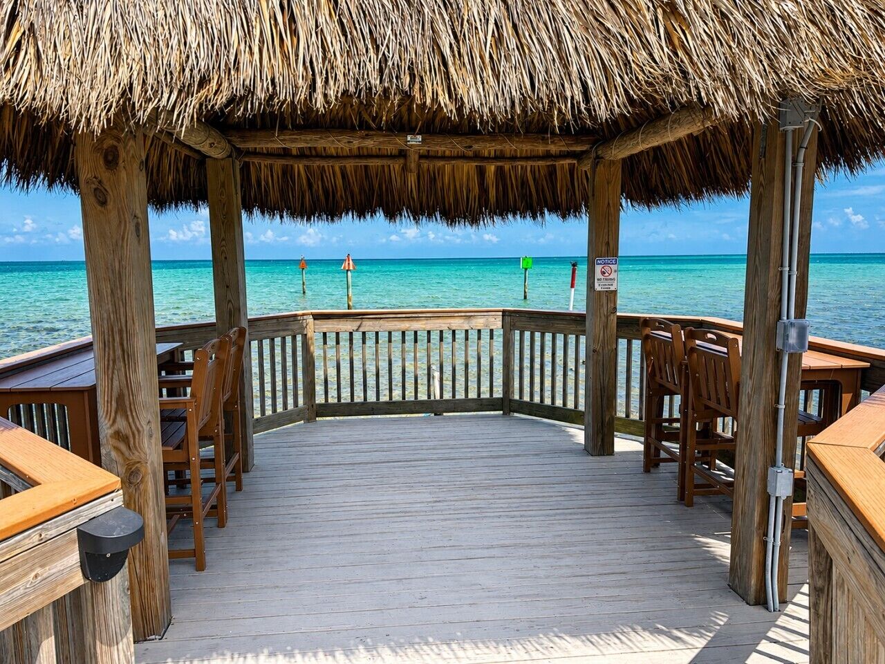 A wooden deck with a thatched roof, wooden chairs, and tables, overlooking a clear blue sea.