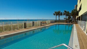 En klarblå pool med en djupmarkering som visar 90 cm, omgiven av ett vitt staket och palmer, med utsikt över havet i bakgrunden.