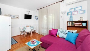 Una sala moderna con un sofá rosa, una mesita con una cesta, un televisor blanco y una estantería con libros y objetos decorativos.