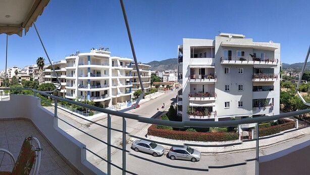 A balcony with a glass table and chairs, overlooking a street with cars and buildings. - Thalia Apartment Kalamata (Kalamata)