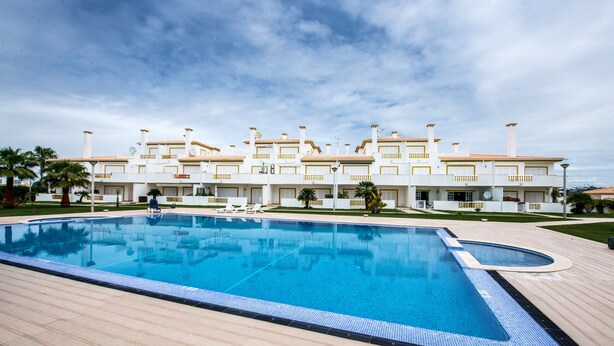 Una gran piscina con terraza de madera, rodeada por un edificio blanco con varios balcones y un cielo azul despejado.