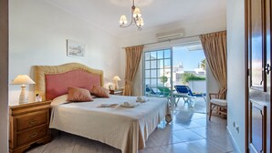 Habitación de hotel con una cama grande, dos mesitas de noche con lámparas, una araña y un balcón con sillas, ofreciendo vistas de la ciudad.