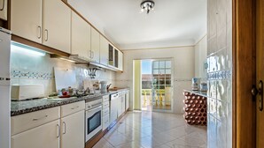 Una cocina con armarios blancos, una estufa y un lavamanos. Se ve un edificio a través de una ventana.