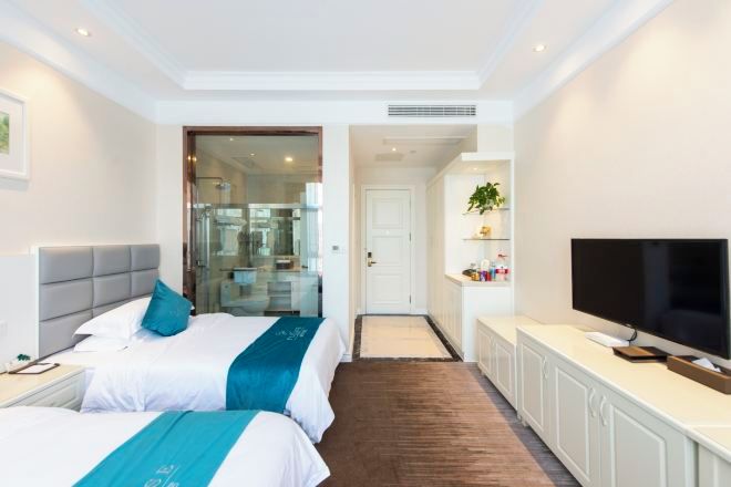 Habitación de hotel con dos camas, televisor, una kitchenette pequeña y un baño visible a través de una puerta de cristal.