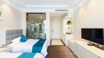 Habitación de hotel con dos camas, televisor, una kitchenette pequeña y un baño visible a través de una puerta de cristal.