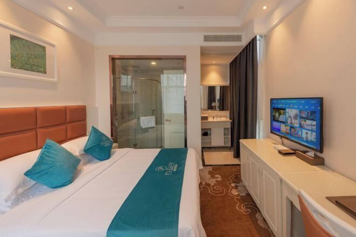 A hotel room with a bed, a TV, a desk, and a bathroom visible through a glass door.