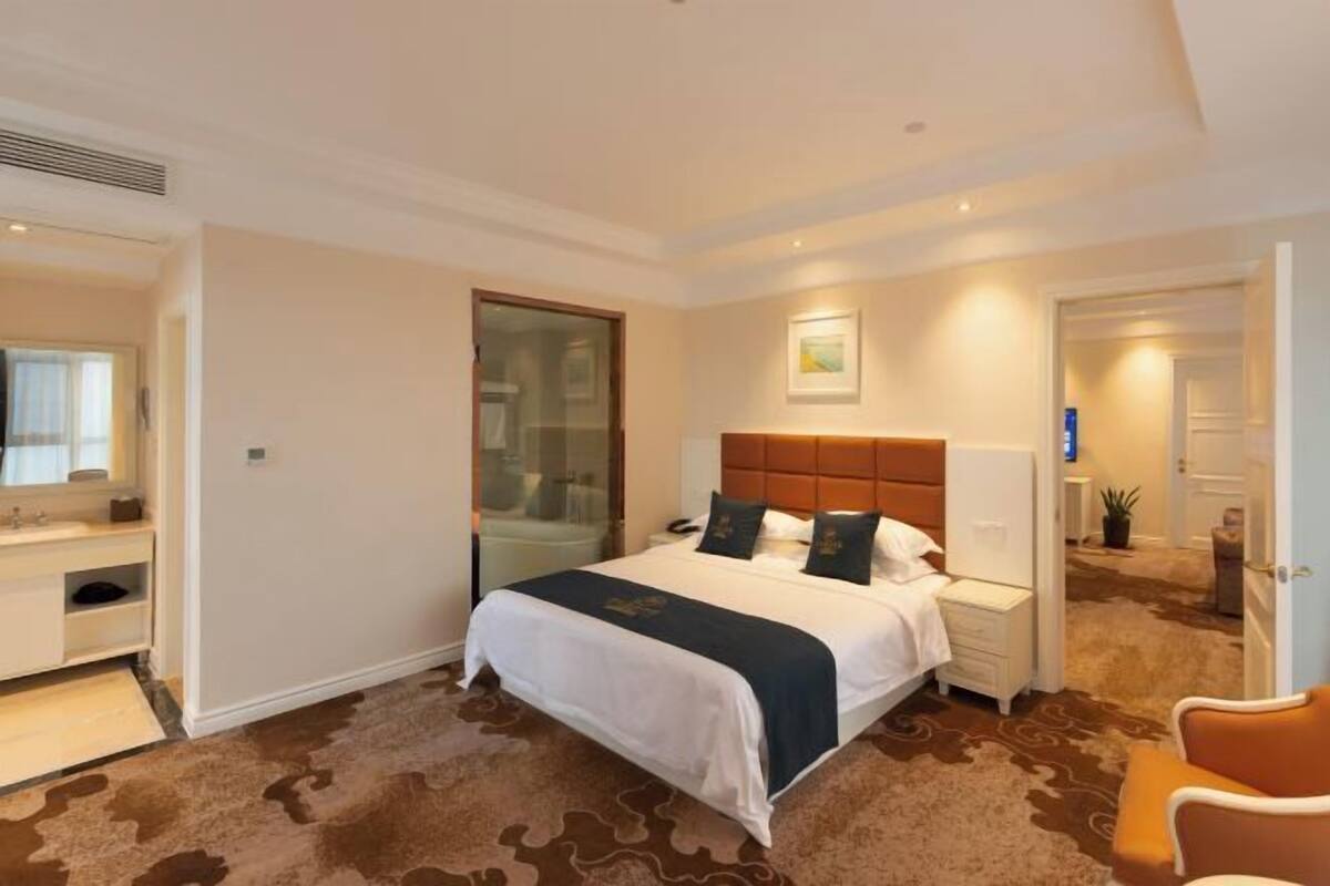 A hotel room with a large bed, a bedside table, a nightstand, a chair, a bathroom with a mirror, and a door leading to another room.