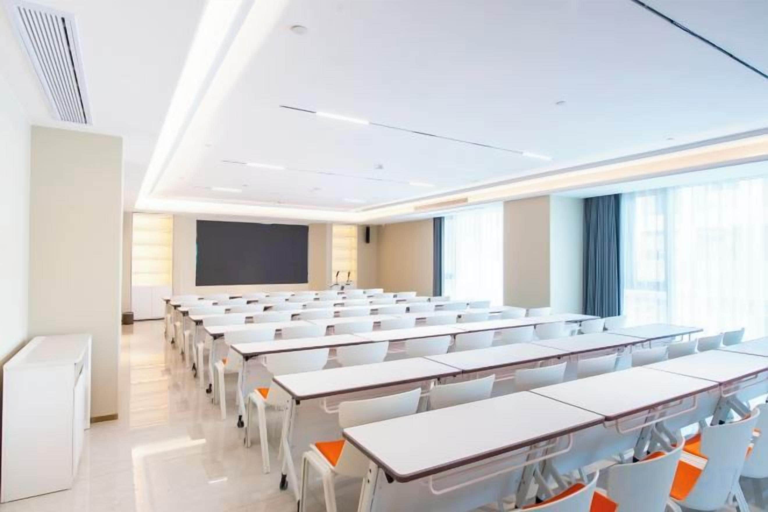 Une salle de classe avec des rangées de bureaux, un écran de projection et une grande fenêtre.