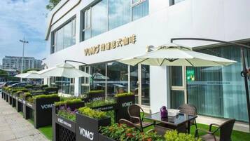 Et udendørs siddeområde med parasoller, stole og plantekasser med teksten 'VOMO' foran en bygning med store vinduer.