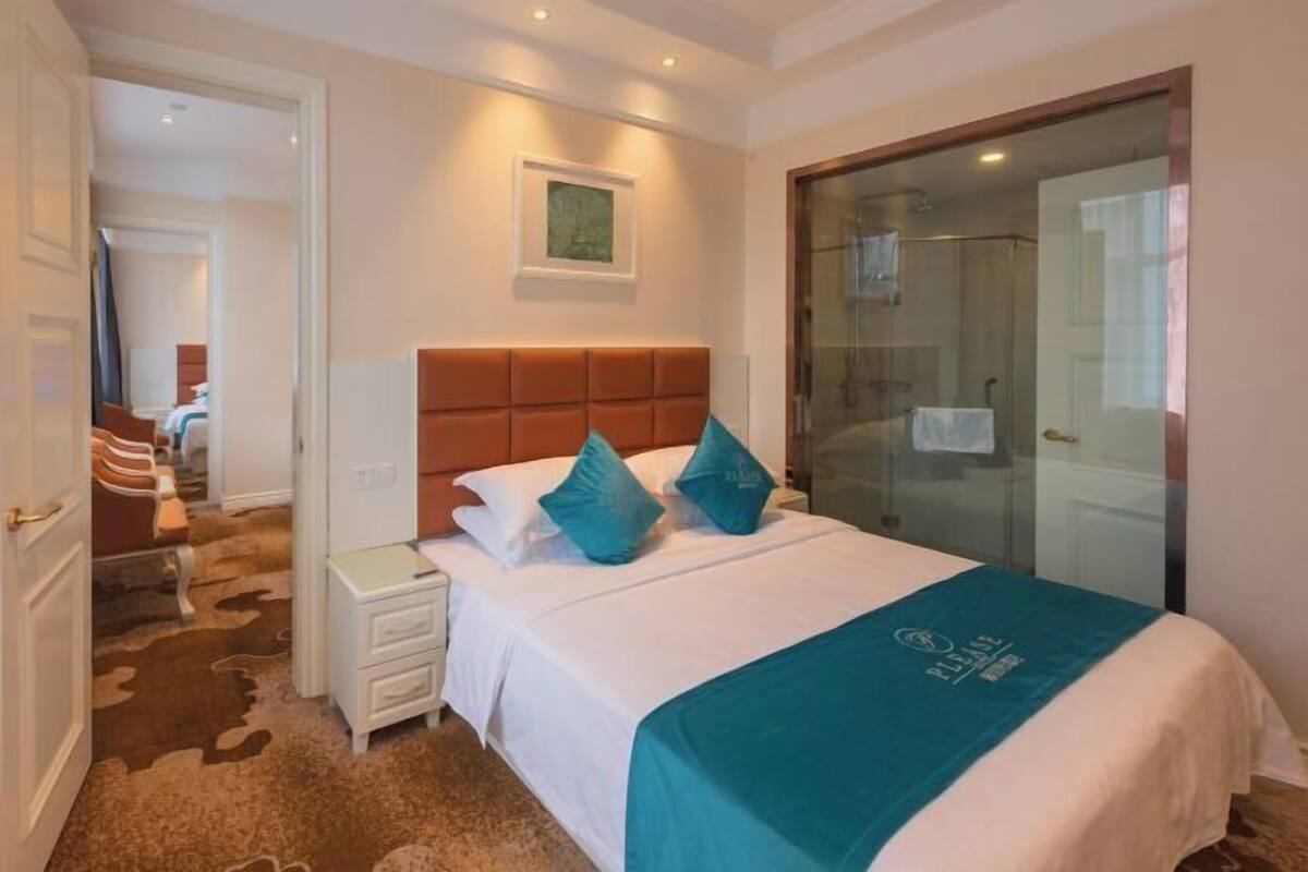 A hotel room with a bed, a nightstand, a glass shower enclosure, and a door leading to another room.