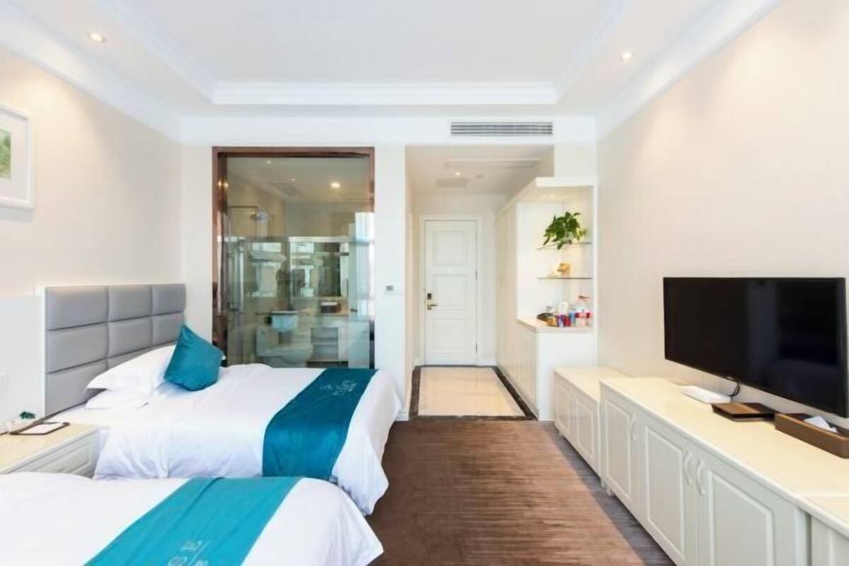 A hotel room with two beds, a TV, a small kitchenette, and a bathroom visible through a glass door.