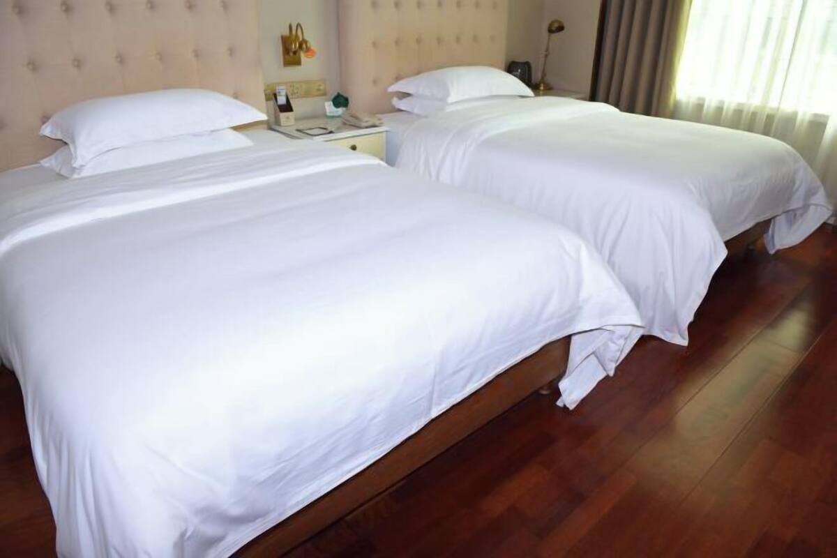 Two single beds with white linens, wooden flooring, a nightstand with a lamp, and a window with curtains.