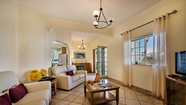 Un salón con sofá, mesa de centro y televisor. Se ve una cocina al fondo, con ventana y cortinas.