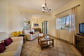 A living room with a sofa, coffee table, and TV. There is a kitchen in the background with a window and curtains.