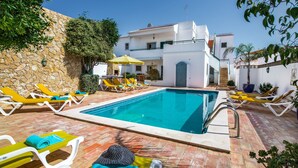 Una piscina con sillas de descanso y sombrillas, rodeada por un muro de piedra y un edificio blanco.