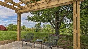 Une terrasse couverte avec deux chaises en métal et une petite table, entourée d’une pergola en bois, le tout se fondant dans un décor verdoyant et luxuriant.