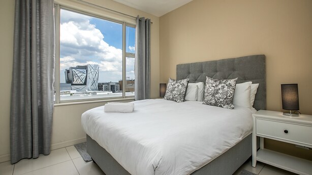 A modern bedroom with a large bed, grey curtains, a view of the cityscape, and a grey headboard. - Designer Apartment in The Iconic Tyrwhitt (Johannesburg)